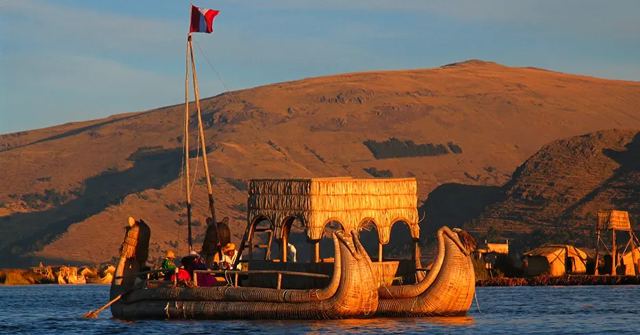 Lugares para visitar en Puno Perú: Explora lo Mejor con Nosotros 1 Personas locales navegando un barco hecho de totora, se puede observar la bandera de Perú en la parte de arriba del barco, con el fondo de una gran montaña y diferentes islas hechas de totora
