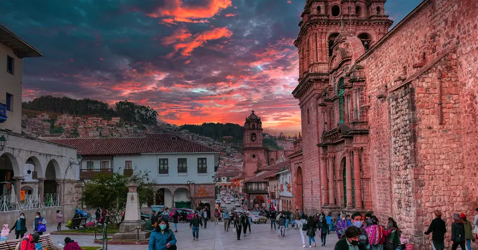 Vista del cusco, iglesia de la merced y al fondo la compañía de Jesús, bastantes personas caminando y un cielo despejado en el atardecer