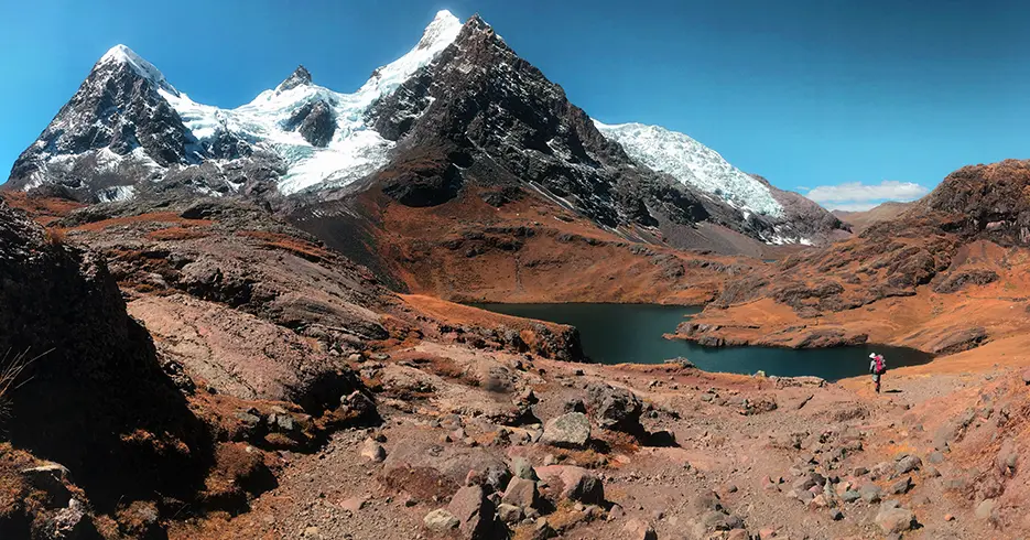 Cómo Realizar el increíble Tour 7 Lagunas de Ausangate en Cusco en 1 Día 2 Nevado ausangate de fondo con una de las 7 lagunas del recorrido, montañas puntiagudas, cielo celeste con una vista de un camino de tierra y piedras y una persona observando la laguna y el nevado.