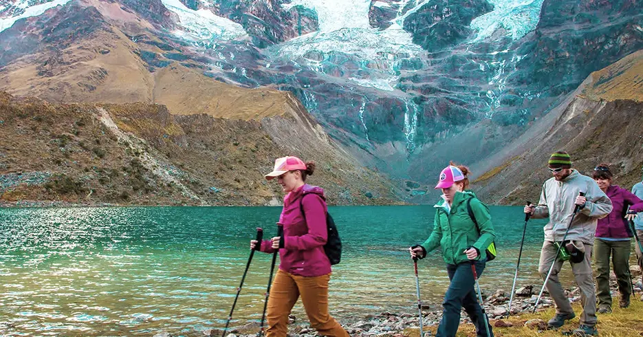 Los 10 Mejores Lugares Turísticos a Visitar en Cusco Perú 2 Turistas en un trekking con sus bastones en la laguna humantay, agua cristalina y turquesa y el nevado humantay en la parte de arriba