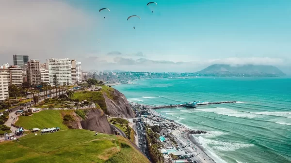 La capital de Perú, Lima Miraflores, se puede ver el océano pacífico 3 paracaidistas en el cielo, un cielo despejado celeste, varis carros y personas.