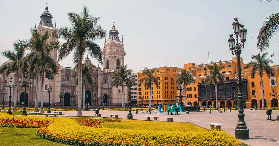 Haz Realidad Tu Viaje a Perú en 7 Días 1 Plaza de armas de lima, con varias palmeras, asientos y una pileta, con la catedral de fondo y al lado el palacio municipal de lima.