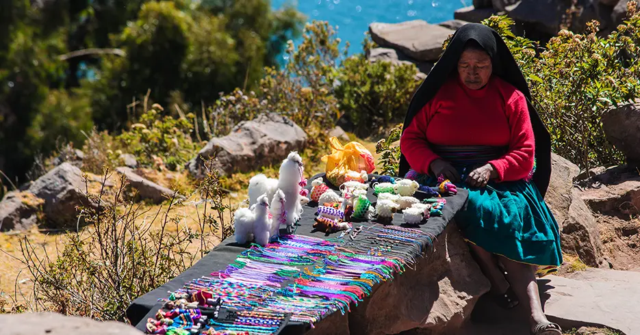 Lugares para visitar en Puno Perú: Explora lo Mejor con Nosotros 3 Persona local en la isla de taquile, tejiendo sus recuerdos para los turistas, de alpacas, pulseras, al fondo se ve el lago Titicaca difuminado y una vista de rocas y plantas verdes