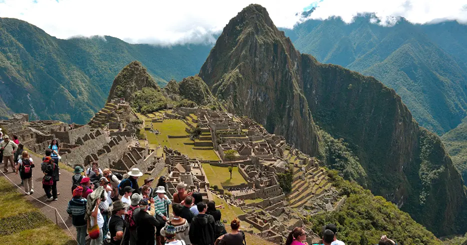 Machu Picchu en la temporada alta, con mucha cantidad de turistas en los caminos de Machu Picchu para ver la maravilla, cielo con muchas nubes, y la vegetación verde