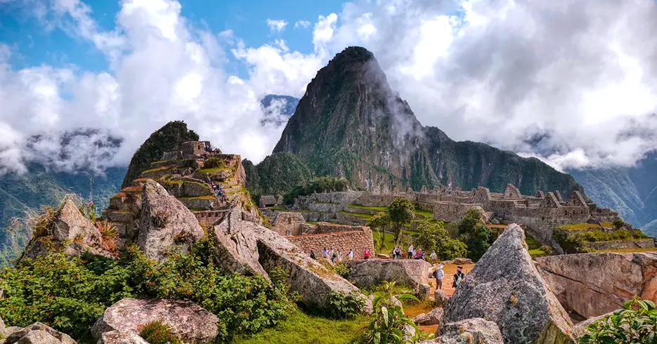 Viaje a los Andes en 11 días: Lugares impresionantes que debes conocer 3 Machu Picchu en cusco, con varias personas, se ven varias piedras y pasto verde y los espacios arqueológicos de fondo en la parte baja, un cielo con varias nubes y una parte el cielo azul