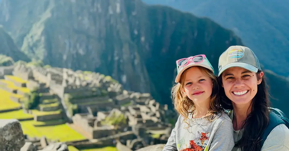 Turistas madre e hija en Machu Picchu felices, las dos con gorra y cabello largo, al fondo Machu Picchu difuminado y las montañas verdes.