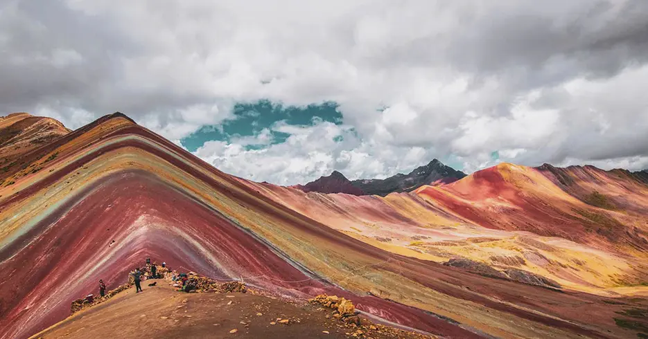 Viaje a los Andes en 11 días: Lugares impresionantes que debes conocer 5 La montaña de los 7 colores en un viaje a los Andes, se ven varios colores, como el rojo, amarillo, naranja y blanco, con un cielo un poco nublado y varias personas en la parte izquierda