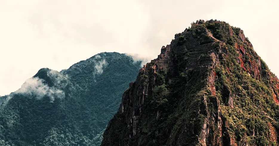 Montaña huaynapicchu en Machu Picchu con un cielo lleno de nubes y al fondo una montaña verde con bastantes árboles.