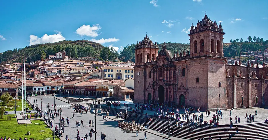 Tour cusco y Machu Picchu 3 Días: Un Viaje Inolvidable a la Ciudad Perdida 1 Vista de la catedral de cusco con bastantes personas en la parte baja, en las escaleras y en la calle, árboles verdes al fondo y un cielo celeste