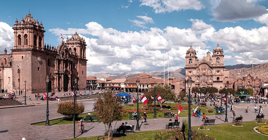 Plaza de armas de cusco con un cielo celeste y bastantes nubes, se puede observar dos inglesias, la catedral y la compañía de Jesús y muchas banderas de Perú