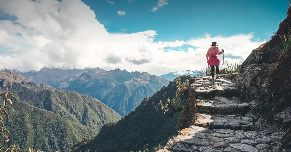Mujer en un trekking en Perú por el camino inca con un camino empedrado, con bastones y vista de grandes montañas con un cielo celeste