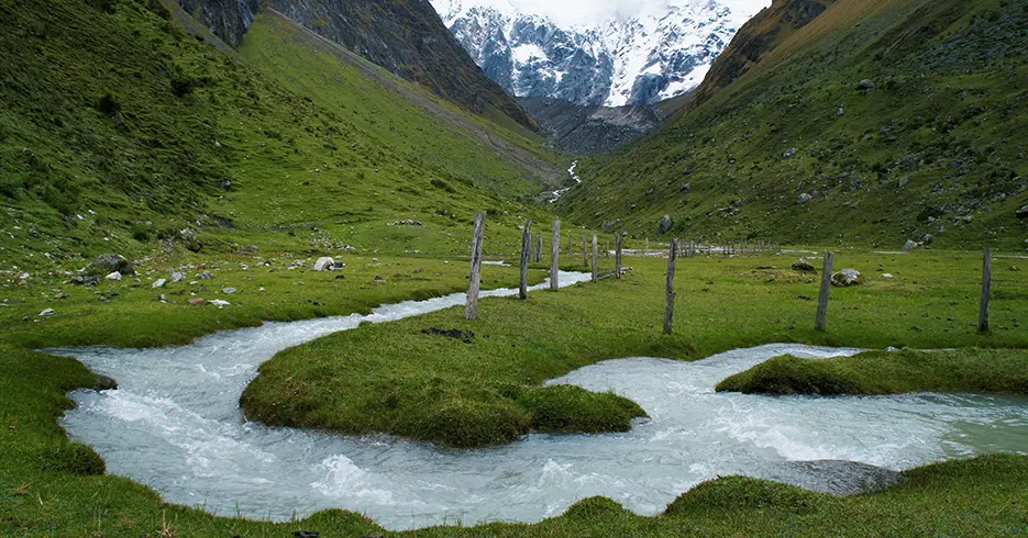 Ruta de trekking en Perú, camino del salkantay, con el nevado al fondo y paisaje de color verde y un pequeño rio