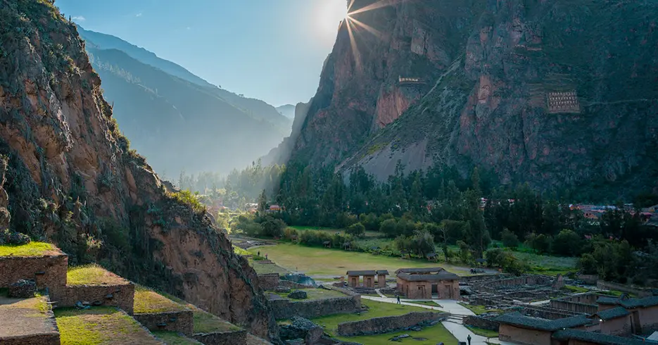 Haz Realidad Tu Viaje a Perú en 7 Días 3 Centro arqueológico de ollantaytambo, en la parte baja se encuentran espacios incas y una vista llena de color verde, y se ve el sol tapado por la montaña de al frente.