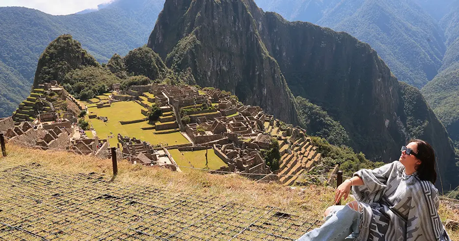 ¿Qué Ver en Perú 9 Días? Los Imperdibles del País 4 Turista en Machu Picchu sentada, posando para la foto y al fondo en la parte baja se ven los centros arqueológicos con la hermosa vista de las montañas y una vegetación verde