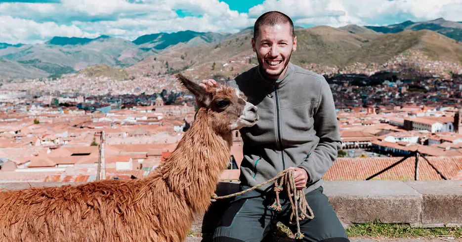 Turista sonriendo con una llama en el mirador de san Cristóbal, al fondo podemos ver la ciudad de cusco y rodeado de montañas