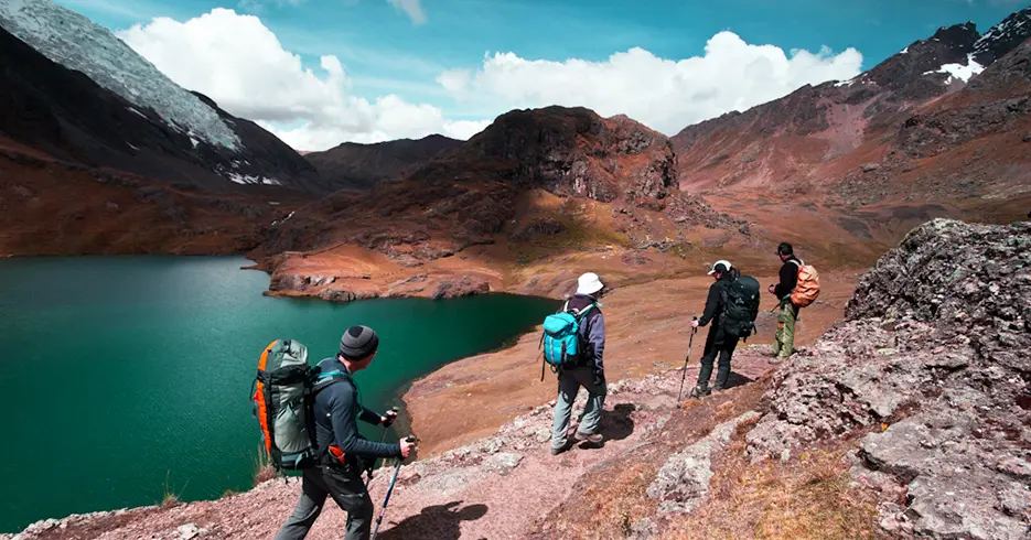 Cómo Realizar el increíble Tour 7 Lagunas de Ausangate en Cusco en 1 Día 3 Turistas caminando con ayuda de bastones de trekking en la ruta de 7 lagunas de ausangate. Cielo despejado celeste, laguna con un color verde- turquesa camino de tierra y piedras