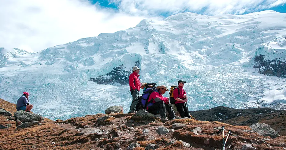 Cómo Realizar el increíble Tour 7 Lagunas de Ausangate en Cusco en 1 Día 4 Turistas descansando en los pies del nevado ausangate con sus bastones de trekking, dos de ellos viendo el paisaje, otros dos descansando, uno sentado alejado de los otros, un camino lleno de piedras y un cielo lleno de nubes blancas.