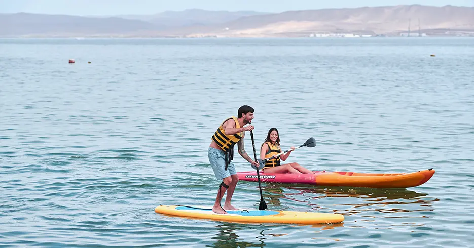 Qué Hacer en Paracas Perú: Experiencias que No Te Puedes Perder 5 Dos turistas haciendo los deportes de kayac y paddle surf con ayuda de los remos en las aguas de paracas. Un bote pequeño rojo con naraja, y una tabla amarilla con azul y chalecos amarillos y rayas negras.