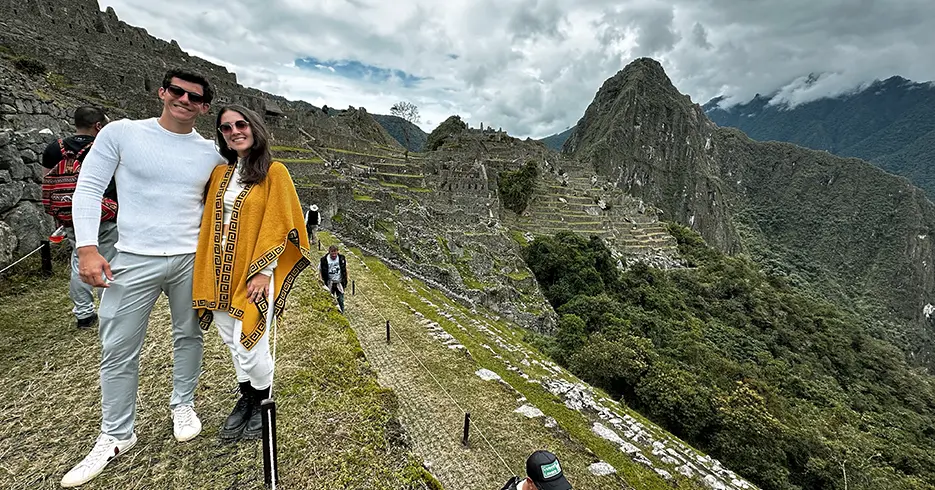 ¿Qué ropa llevar a Machu Picchu? La guía definitiva para viajeros 2 Pareja de turistas sonriendo en Machu Picchu, caminando por los Andes y vista de las construcciones incas y las montañas y los árboles